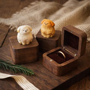 Könnte beinhalten: Drei quadratische Ringkästchen aus dunklem Holz mit Klappdeckeln. Zwei Kästchen sind geschlossen und mit kleinen, handgeschnitzten Hunde-Holzfiguren in Creme und Goldbraun verziert. Das dritte Kästchen ist geöffnet und zeigt ein dunkles Samtfutter und einen Goldring.