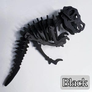 May include: A black dinosaur skeleton puzzle. The puzzle is made of textured material and features a detailed design of a Tyrannosaurus Rex. The word "Black" is in a white box at the bottom right.