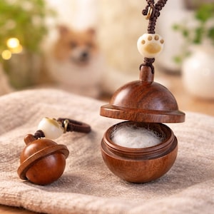 Op de afbeelding: Een houten dierenurn met een pootafdrukbedel. De urn is open en onthult een witte vulling. Een kleinere bijpassende bedel is ook zichtbaar. De urn en de bedel zijn bruin met een lichtgekleurde pootafdrukaccent.