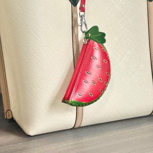 K&ouml;nnte beinhalten: Eine rote, lederne Geldb&ouml;rse in Wassermelonenform mit einem gr&uuml;nen Blattakzent und einem roten Rei&szlig;verschluss. Die Geldb&ouml;rse ist an einer wei&szlig;en Tragetasche mit rotem Riemen und silbernem Verschluss befestigt.