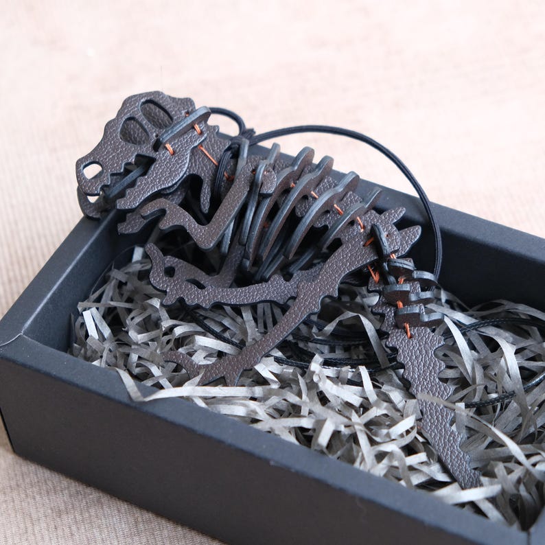 May include: A dark brown leather dinosaur skeleton pendant necklace. The pendant is intricately cut and stitched with orange thread, presented in a black gift box filled with shredded paper.