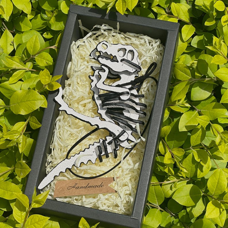 May include: A white and black dinosaur skeleton cut-out in a black box filled with shredded paper. The dinosaur is made of layered materials and has black accents. A small brown tag with the word "Handmade" is attached to the box.