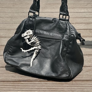 May include: Black leather handbag with double handles and a zipper pocket. A white and black dinosaur skeleton charm hangs from the handle. The bag is sitting on a wooden surface.