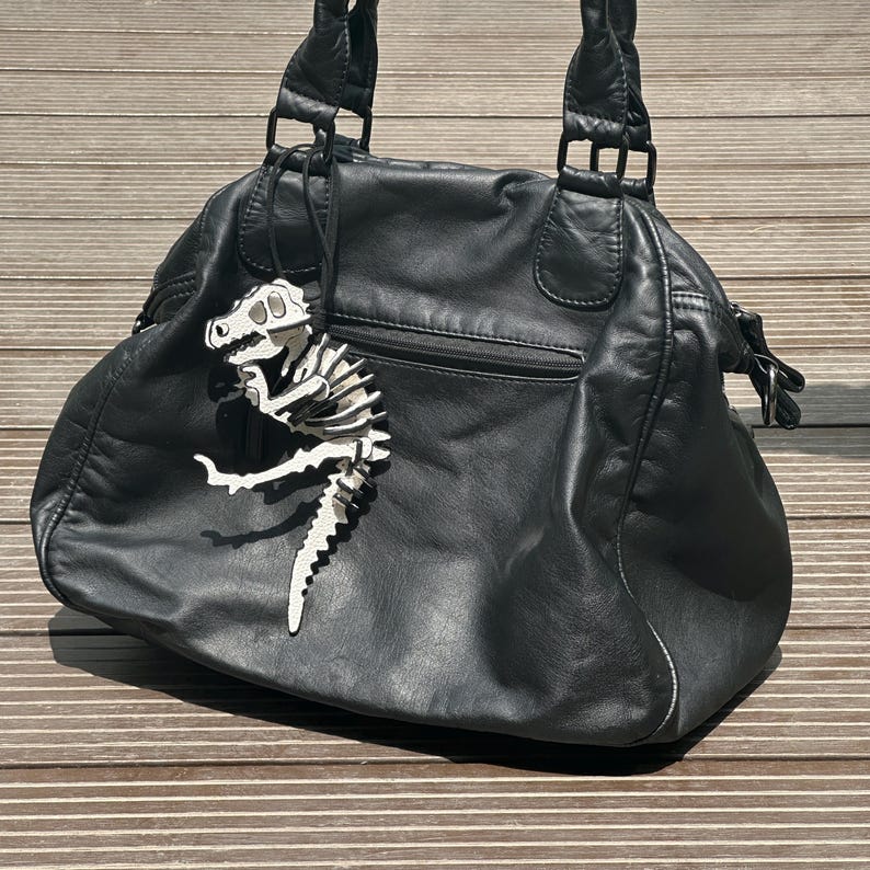 May include: Black leather handbag with double handles and a zipper pocket. A white and black dinosaur skeleton charm hangs from the handle. The bag is sitting on a wooden surface.
