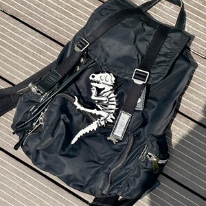 May include: Black backpack with a white dinosaur skeleton patch. The backpack has adjustable straps, a top handle, and multiple zippered compartments. The bag is made of a durable, possibly nylon, material.