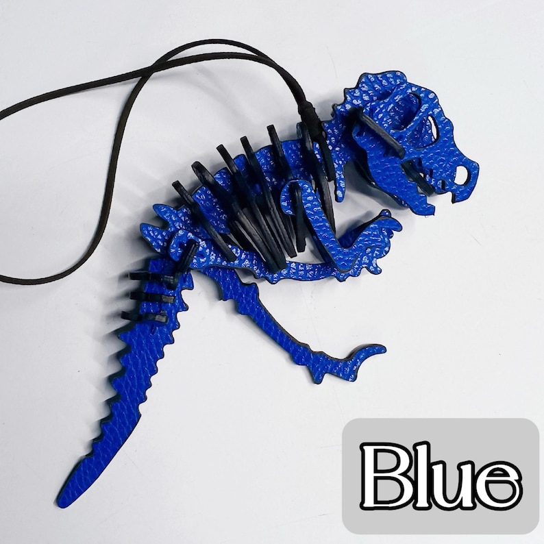 May include: A blue dinosaur skeleton pendant with a black cord. The pendant is made of a textured material and features the word "Blue" in a white font on a gray background. The dinosaur pendant is a unique accessory.