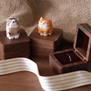 Puede incluir: Figuras de gato de madera en tonos marrón, blanco y naranja sobre cajas de madera oscura. Una caja está abierta, revelando un anillo de oro. Una cinta blanca con rayas marrones en primer plano.