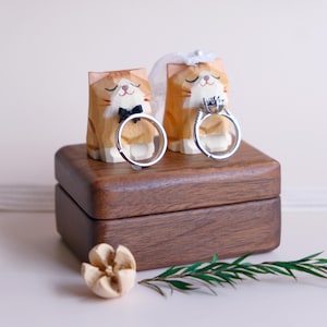 Op de afbeelding: Houten kattenbeeldjes, één met een vlinderdas en de ander met een sluier, die trouwringen vasthouden. De beeldjes staan op een houten doos, met een houten bloem en een takje groen. De ringen zijn zilver.