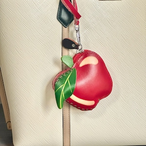 K&ouml;nnte beinhalten: Ein rotes, ledernes, apfelf&ouml;rmiges M&uuml;nzfach mit einem gr&uuml;nen Blatt und einem roten Rei&szlig;verschluss. Das M&uuml;nzfach ist an einer cremefarbenen Tragetasche mit rotem Riemen und silberfarbenen Beschl&auml;gen befestigt.