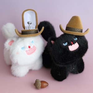 Puede incluir: Dos figuras de gato de peluche, una blanca y otra negra, cada una con un sombrero de vaquero marrón. El gato blanco sostiene un joyero con un anillo de diamantes. Una bellota descansa sobre la superficie rosa.
