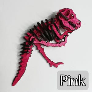 May include: A pink dinosaur skeleton puzzle with black accents. The dinosaur is made of interlocking pieces and has a textured surface. The word "Pink" is in a gray box in the lower right corner.