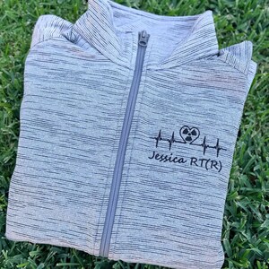 May include: A light gray heathered zip-up jacket with a high collar and a center zipper. The chest features a black embroidered design of a heartbeat line with a radiation symbol inside a heart shape, followed by the text Jessica RT(R).