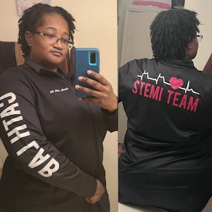 May include: Black zip-up jacket with white text on the sleeve and the words "STEMI TEAM" in red and white on the back. The jacket features a heart and EKG line design. The jacket is worn by a person.