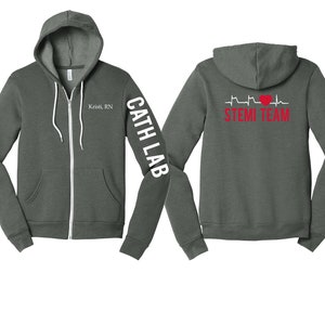 May include: A dark gray zip-up hoodie with a white heart and EKG graphic on the back that says "STEMI TEAM". The left sleeve has white text that says "CATH LAB". The front of the hoodie has white text that says "Kristi, RN".
