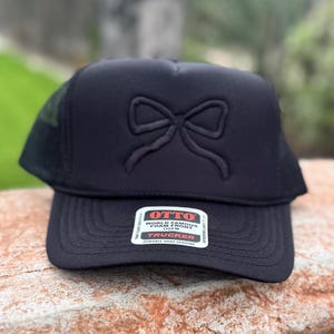May include: Black trucker hat with a bow design on the front. The hat has a mesh back and a curved brim. The hat has a tag that says "OTTO WORLD FAMOUS FOAM FRONT TRUCKER".