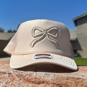 May include: A beige trucker hat with a white embroidered bow on the front. The hat has a mesh back and a white adjustable strap. The hat has a label that says "Otto World Famous Trucker Rucker Durable Snap Closing".