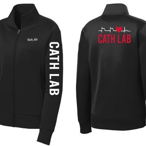 May include: Black zip-up jacket with white text on the sleeve that reads "Cath Lab" and white text on the front that reads "Kidly, RN". The back of the jacket has a white EKG line with a red heart and the text "Cath Lab" in white.