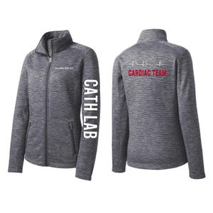 Peut inclure: Veste grise zippée avec le texte "CATH LAB" sur la manche gauche et "CARDIAC TEAM" au dos. La veste a un col montant, deux poches avant et une fermeture éclair. Le dos présente un graphique de moniteur de fréquence cardiaque.