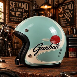 Puede incluir: Un casco de moto abierto de color azul claro con una franja blanca curva, un gráfico circular rojo y el texto negro "Ganbate" junto con caracteres japoneses. El casco tiene un borde negro, broches de metal plateado y una correa para la barbilla.