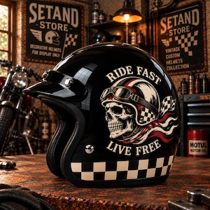 Puede incluir: Un casco de moto abierto de color negro brillante con un gráfico de una calavera que lleva gafas y casco, junto al texto RIDE FAST LIVE FREE. El casco incluye una visera negra y un patrón de cuadros blancos y negros en el borde inferior.