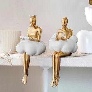 May include: Two gold-coloured figurines of seated figures with cloud-shaped bases. One holds a book, the other a sphere. The figures are sitting on a white shelf. The cloud bases are white, and the figures are gold.