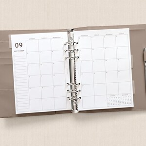 May include: A taupe-colored six-ring binder planner open to a September monthly calendar view. The white pages feature a grid layout with the number 09 and the word SEPTEMBER printed at the top left. A silver metal pen is tucked into the side loop.