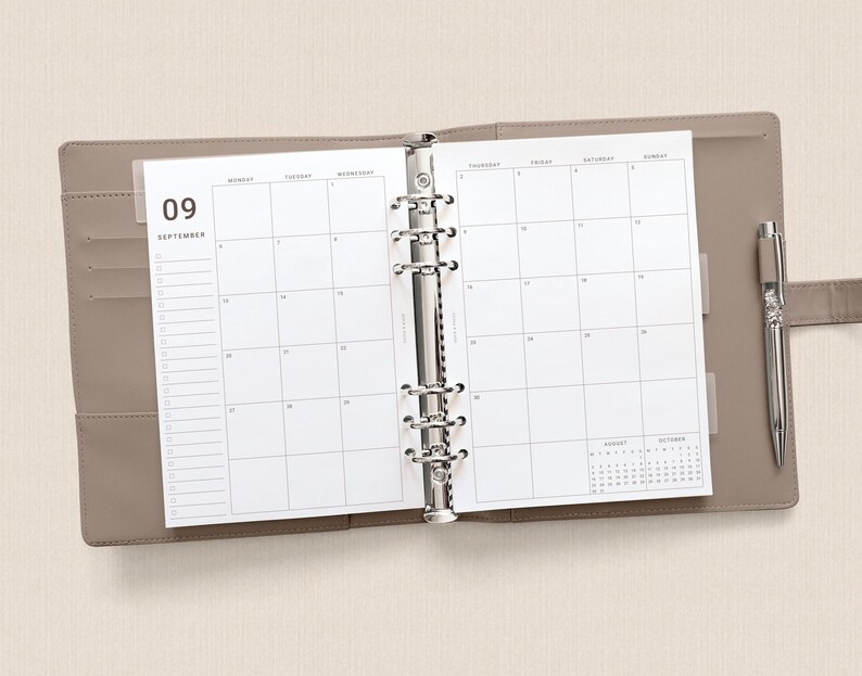 May include: A taupe-colored six-ring binder planner open to a September monthly calendar view. The white pages feature a grid layout with the number 09 and the word SEPTEMBER printed at the top left. A silver metal pen is tucked into the side loop.