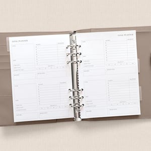 May include: Open planner with "GOAL PLANNER" text on the top of the pages. The planner is open to a spread with sections for goals, actions, and reviews. A silver pen rests on the right side of the planner, which is a light brown color.