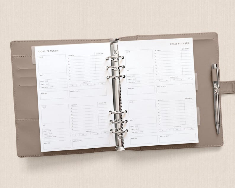 May include: Open planner with "GOAL PLANNER" text on the top of the pages. The planner is open to a spread with sections for goals, actions, and reviews. A silver pen rests on the right side of the planner, which is a light brown color.