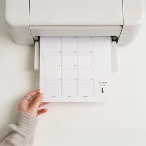 May include: A white printer outputting a paper sheet featuring a January 2027 calendar grid. The calendar displays the month name, year, and days of the week in a clean, black sans-serif font. The grid layout includes numbered dates from 4 to 27.
