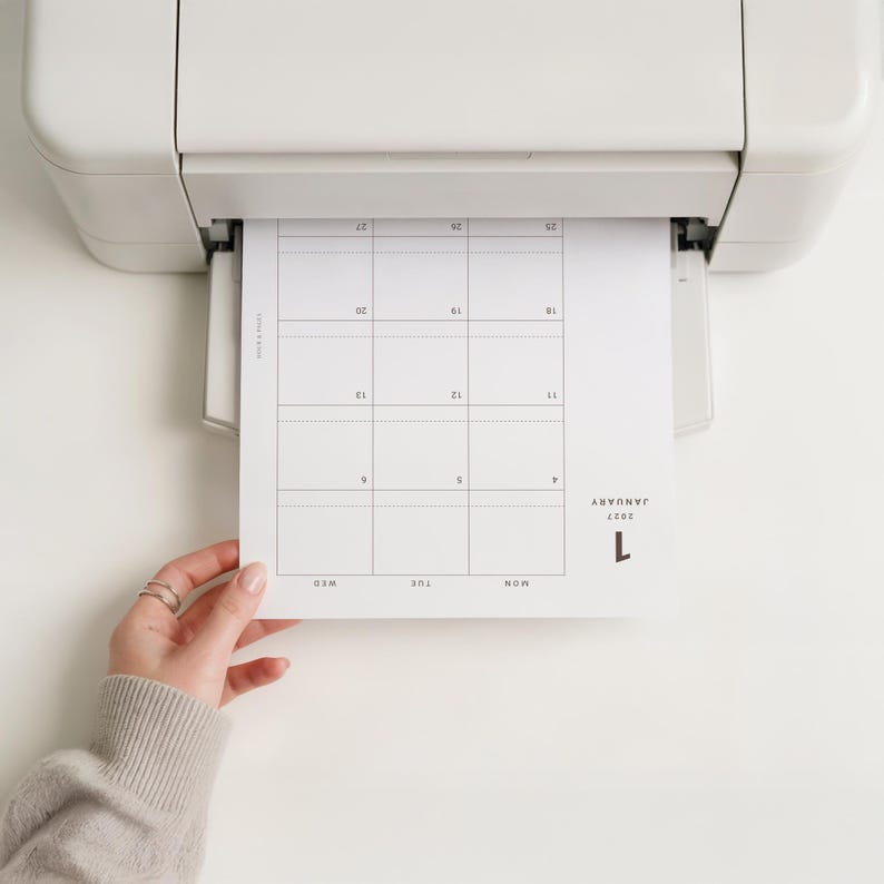 May include: A white printer outputting a paper sheet featuring a January 2027 calendar grid. The calendar displays the month name, year, and days of the week in a clean, black sans-serif font. The grid layout includes numbered dates from 4 to 27.