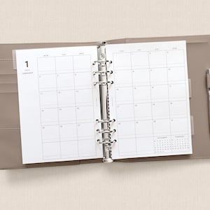 May include: A taupe-colored six-ring binder planner open to a January monthly calendar view. The white paper pages feature a grid layout with the text 1 JANUARY. A silver metal pen is held in a loop on the right side of the binder cover.