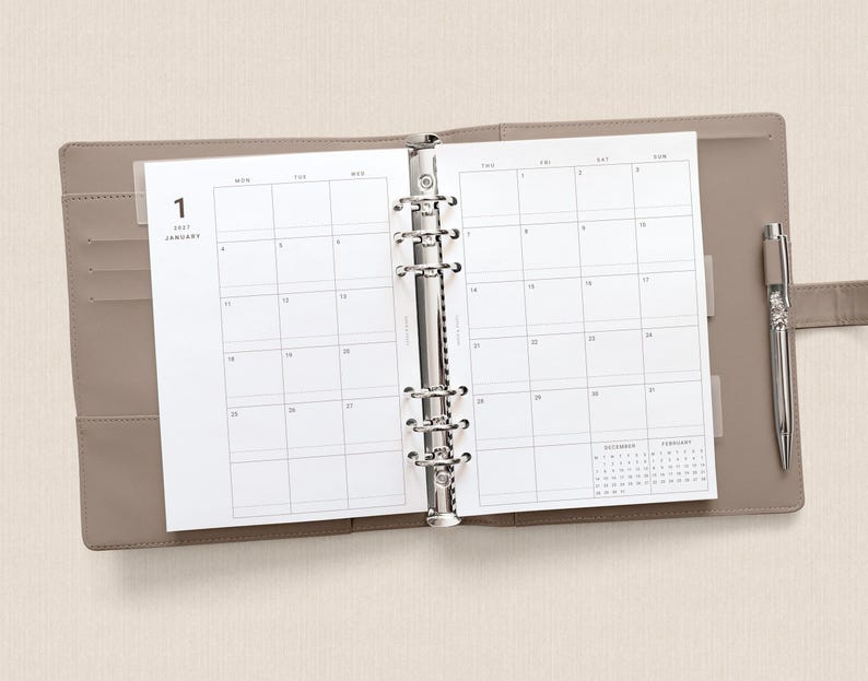 May include: A taupe-colored six-ring binder planner open to a January monthly calendar view. The white paper pages feature a grid layout with the text 1 JANUARY. A silver metal pen is held in a loop on the right side of the binder cover.
