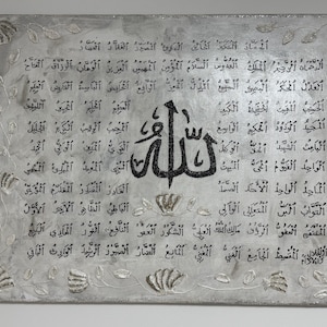 May include: A silver-toned canvas art piece featuring Arabic calligraphy in black. The artwork is adorned with floral and leaf-like embellishments, creating a textured and elegant aesthetic. The central focus is a stylized Arabic word.