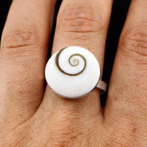 May include: A ring featuring a circular white shell cabochon with a prominent dark brown spiral pattern centred on the surface. The shell is set into a simple metal band that encircles the finger, showcasing a natural organic design element.