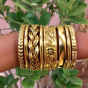 May include: A set of seven gold-toned rings featuring various designs including a chevron pattern, a braided texture, a smooth polished band, and a band with an embossed floral and leaf motif, all displayed together on a finger.
