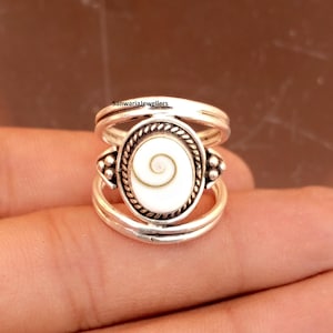 Könnte beinhalten: Ein silberner Ring mit einem ovalen, weißen Mittelstück mit einem Spiraldesign. Der Ring hat ein Doppelband und dekorative Elemente an den Seiten. Der Text "Sanwariajewellers" ist sichtbar.