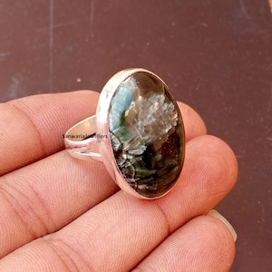 May include: Oval ring with a dark green gemstone and silver setting. The gemstone has a textured appearance with lighter green and silver inclusions. The ring's band is silver. The ring is shown on a hand. The gemstone measures approximately 2 cm in length.