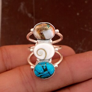 Könnte beinhalten: Drei silberne Ringe mit ovalen Edelsteinen. Der oberste Ring hat einen weißen und goldenen Stein, der mittlere Ring einen weißen Stein mit einem Spiralmuster und der untere Ring einen türkisfarbenen Stein. Die Ringe werden auf einer Hand gezeigt.