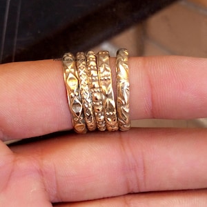 Peut inclure: Cinq bagues en métal doré avec des motifs en relief distincts, incluant des formes géométriques, des croisillons et des textures perlées. Les bagues sont empilées côte à côte et le texte PUSHPALATAJEWELLERS est visible sur la première bague.