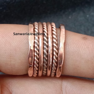 Pode incluir: Um conjunto de seis anéis de cobre com texturas alternadas, incluindo bandas lisas marteladas e bandas trançadas tipo corda. O texto SanwariaJewellers é visível à esquerda. Estas bandas metálicas têm um tom castanho-avermelhado quente.