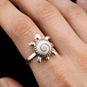 Peut inclure: Une bague en argent avec un motif de tortue incrusté d'une coquille en spirale blanche et brune. La tortue possède une tête et quatre pattes en argent poli, avec la coquille en spirale formant le centre du corps, fixée dans un serti dentelé.