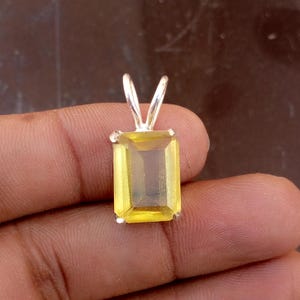 May include: A rectangular, yellow gemstone pendant with a silver bail. The gemstone is a bright, translucent yellow. The pendant is held by a hand against a brown background. The bail is a simple, V-shaped design.