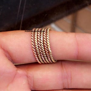 Puede incluir: Un conjunto de cinco anillos de metal fino y trenzado con tonos alternos de oro y oro rosa. Los anillos tienen una apariencia texturizada similar a una cuerda y se usan juntos en un dedo. El texto PUSHPALATAJEWELLERS es visible en el anillo.