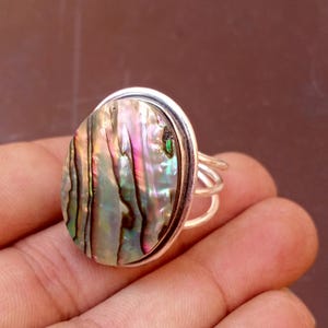 Può includere: Un anello in argento con un grande inserto ovale in conchiglia di abalone. La conchiglia presenta vortici iridescenti di colori rosa, verde e blu con motivi lineari marrone scuro. Il gambo dell'anello è composto da tre fili d'argento paralleli.