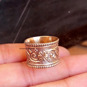 May include: A detailed close-up of a gold-colored ring. The ring features an intricate floral design with a beaded border. The ring's surface is adorned with a pattern of flowers and swirling vines. The ring is a piece of jewelry.