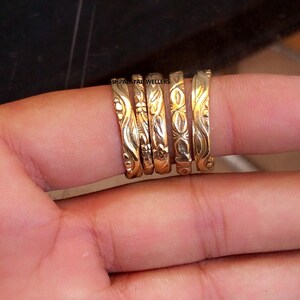 May include: Five gold-toned rings with distinct embossed patterns, including floral, geometric, and swirling designs. The rings are arranged side-by-side, and the text SHPALATAJEWELLERS is visible in small black lettering across the top of the bands.