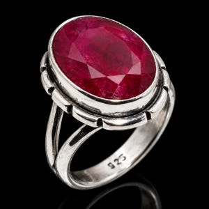 Puede incluir: Un anillo de plata de ley con una gran piedra preciosa de color rojo intenso de corte ovalado engastada en un bisel. La banda de plata tiene un diseño de vástago dividido y está marcada con el número 925 en el interior, indicando la pureza del metal.