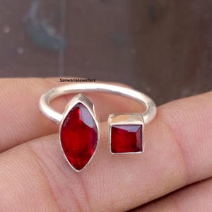May include: A silver ring with two red gemstones. One is a marquise shape, the other a square. The ring has the text "Sanwariajewellers" on the band.