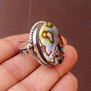 May include: A silver ring featuring an oval abalone shell with iridescent colors of green, blue, and purple. The ring has a detailed silver band with a textured design and the text "SanwariaJewellers".